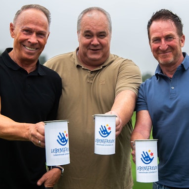 Drei Männer in Poloshirts halten Spendendosen mit der Aufschrift: „Lebensfreude schenken“ in die Kamera.