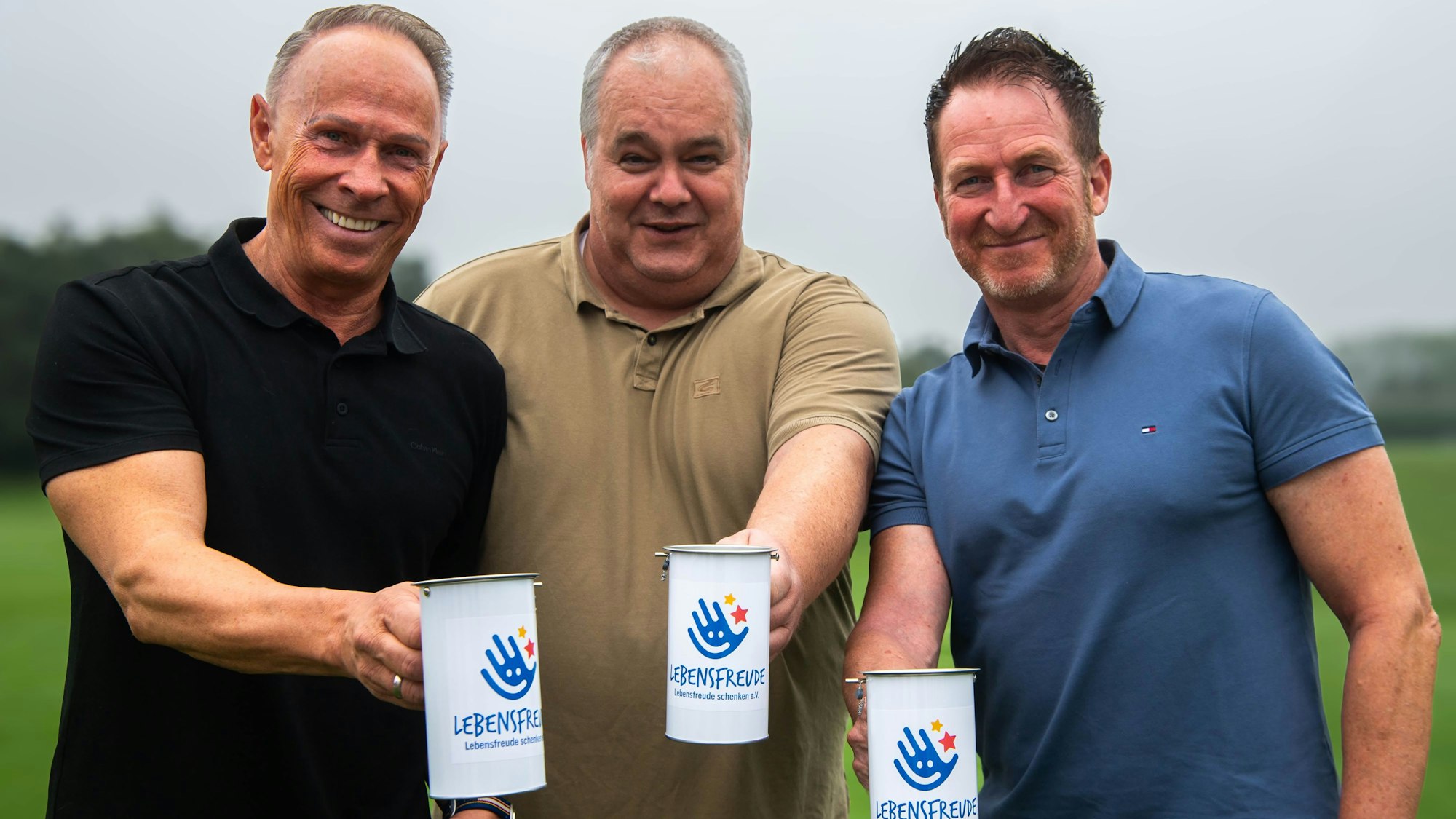 Drei Männer in Poloshirts halten Spendendosen mit der Aufschrift: „Lebensfreude schenken“ in die Kamera.