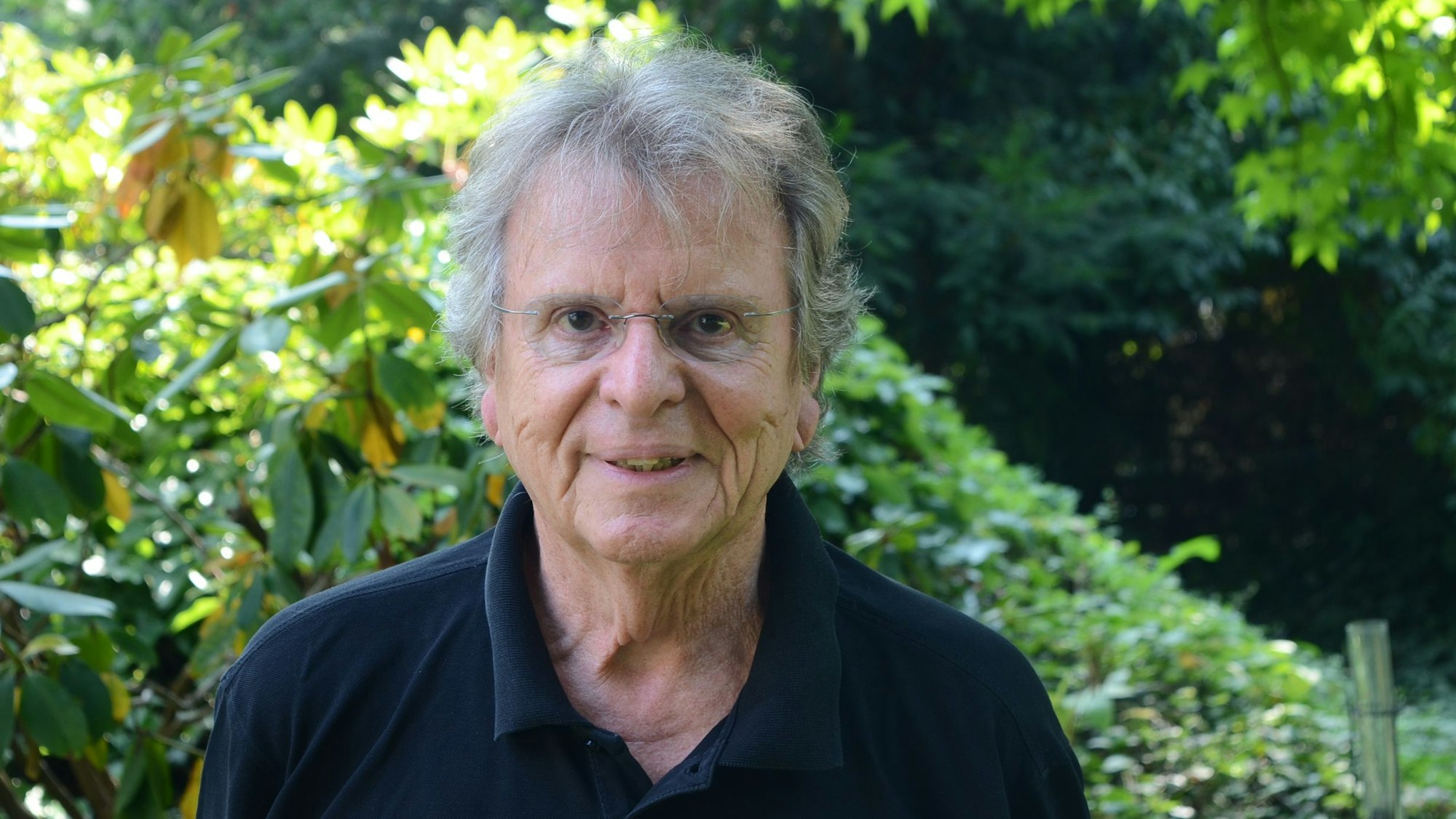 Ein Mann mit Brille und schwarzem Polohemd vor grünen Büschen in einem Garten.