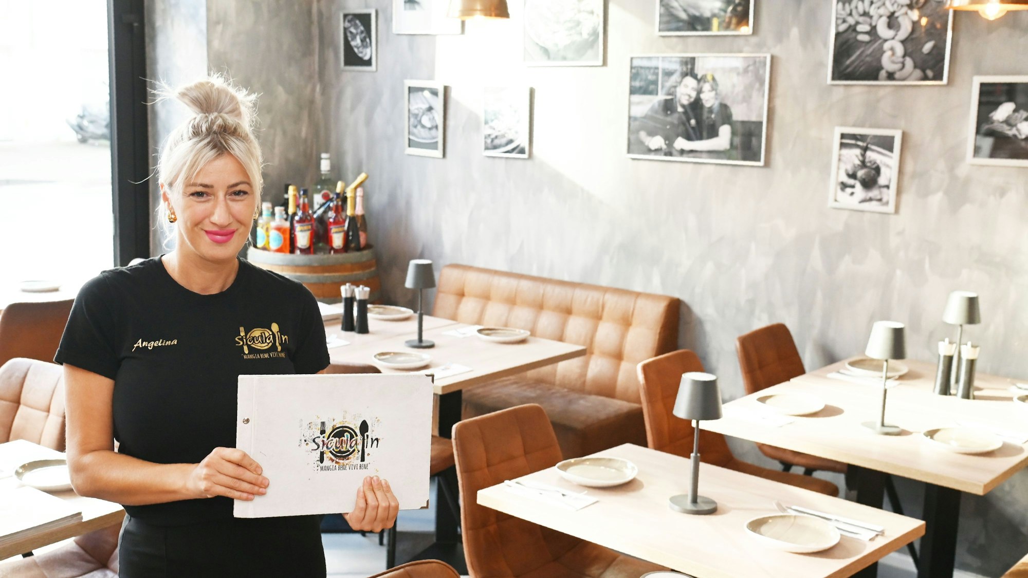 Restaurant-Chefin Angelina zeigt im Gastraum eine Speisekarte und lächelt in die Kamera.