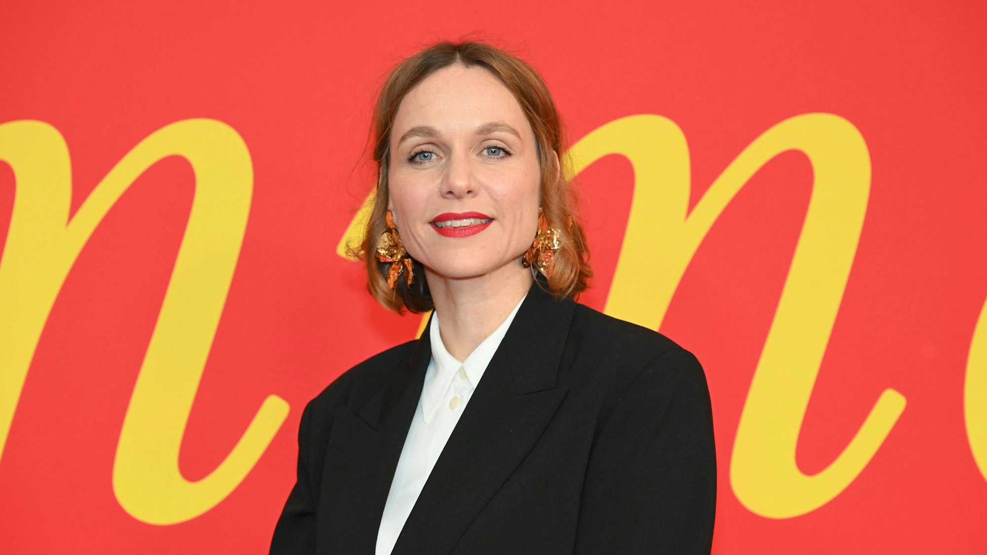 Schauspielerin Nadja Becker steht vor einer roten Wand mit gelber Schrift. Sie trägt eine weiße Bluse und eine schwarze Jacke.