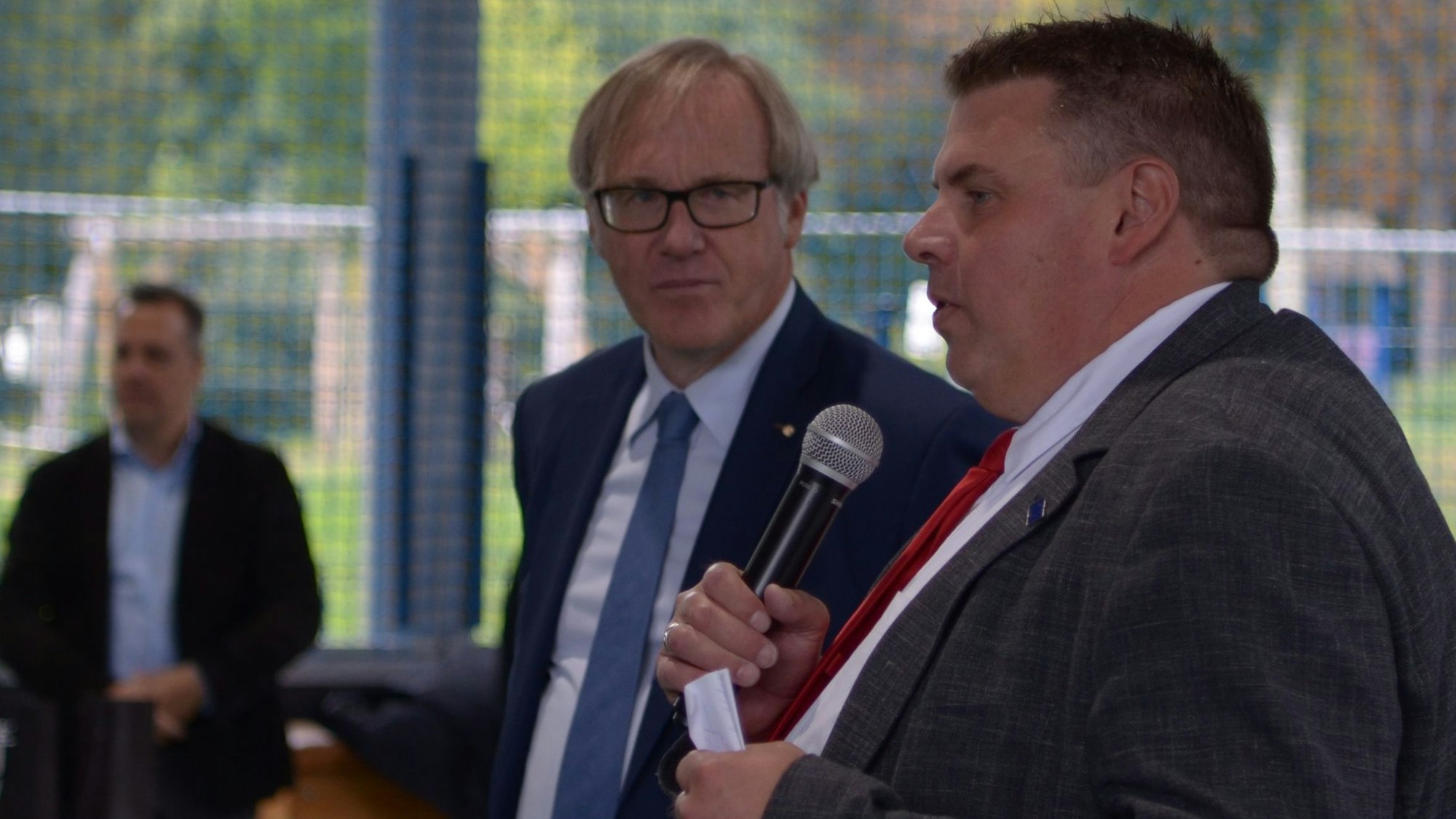 Zwei Männer stehen nebeneinander, einer spricht in ein Mikrofon.