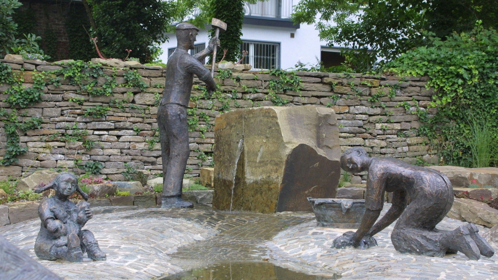 In einer Brunnenlandschaft steht ein Mann mit Hammer, eine Frau putzt und ein Kind spielt.
