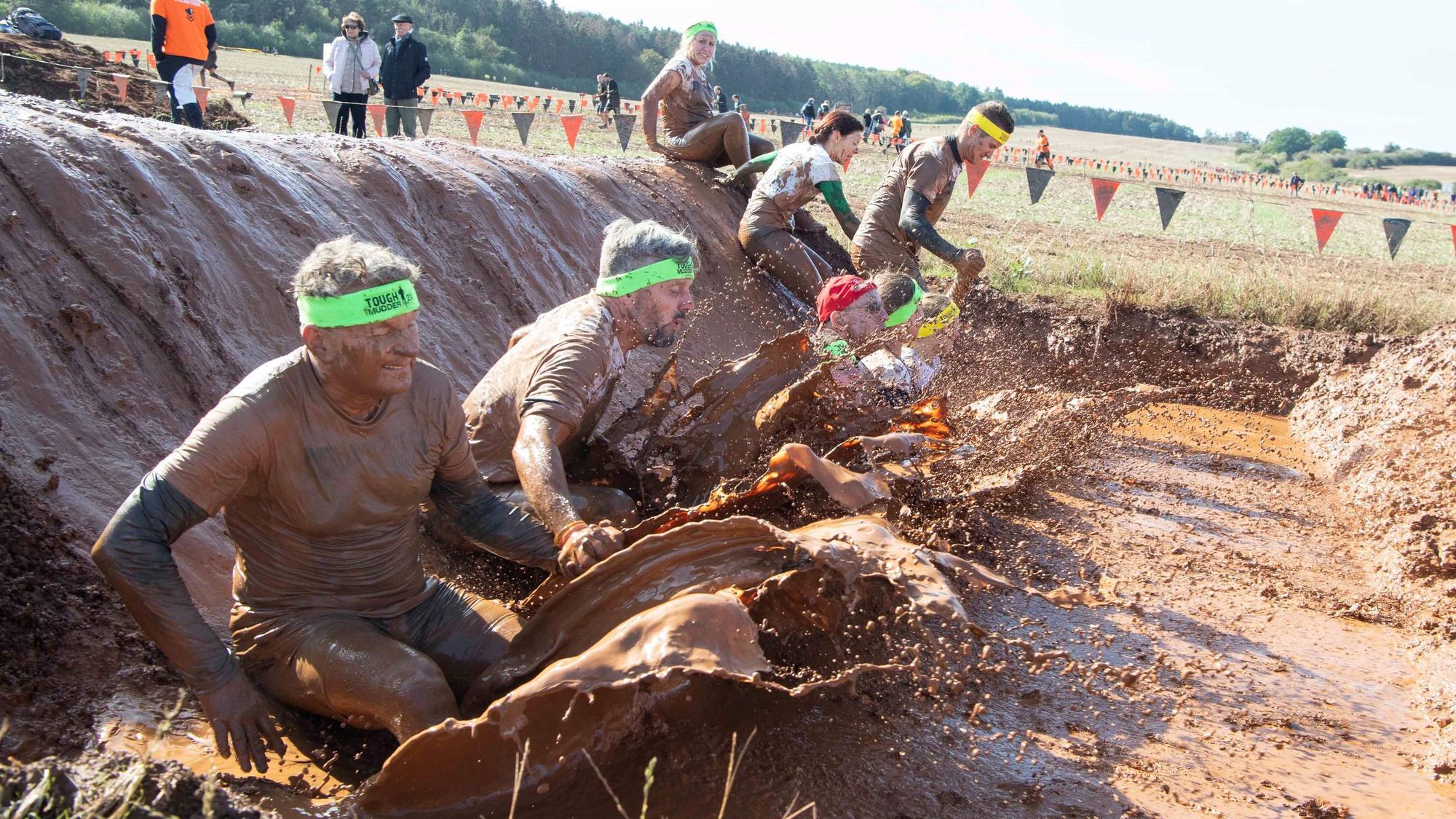 Das Bild zeigt einige Teilnehmer des Tough Mudder, wie sie in einen Schlammgraben rutschen.