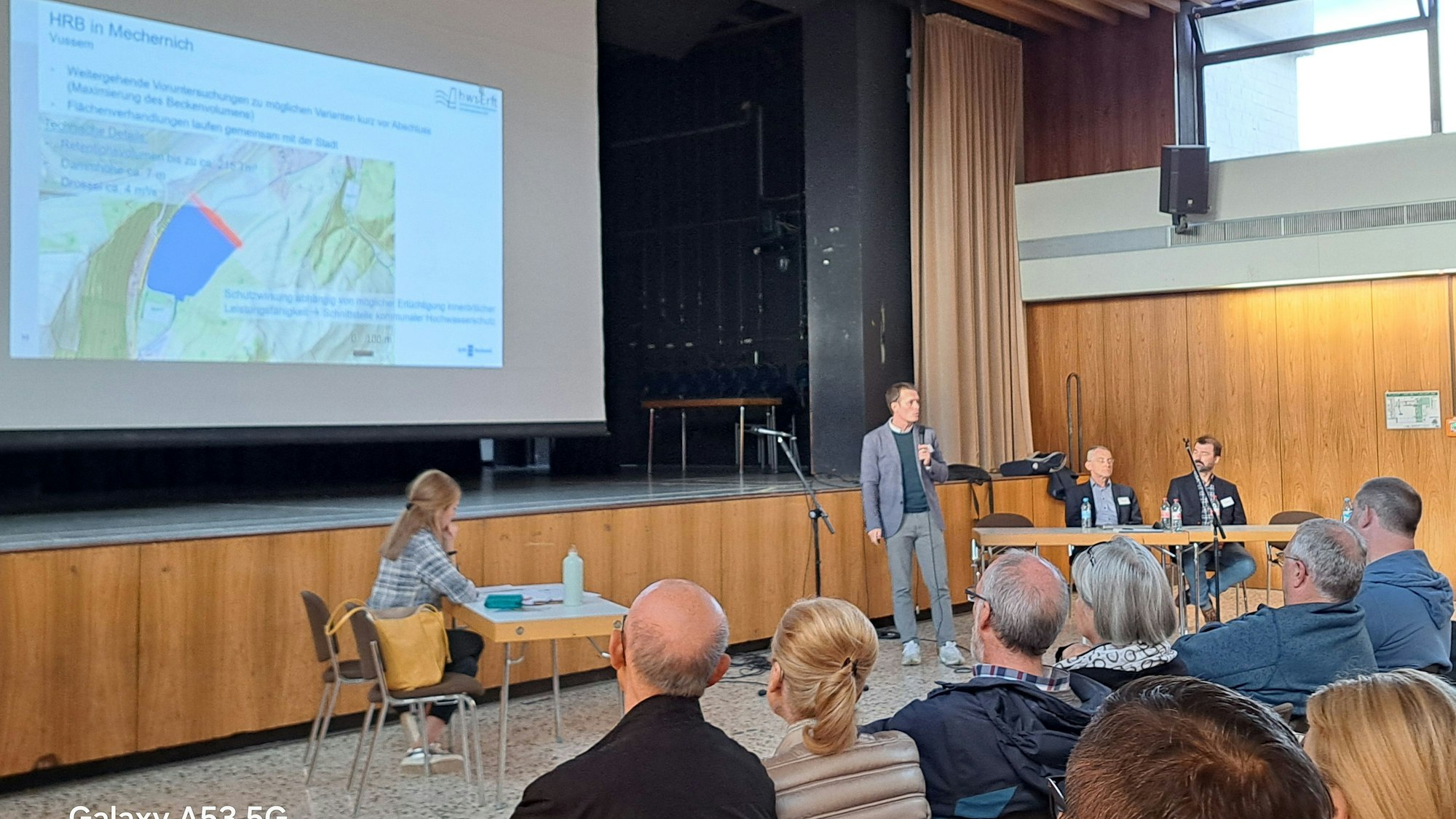 Daniel Bittner steht am Mikrofon, auf einer Leinwand ist die schematische Darstellung eine Rückhaltebeckens zu sehen, Im Vordergrund sitzt das Publikum.