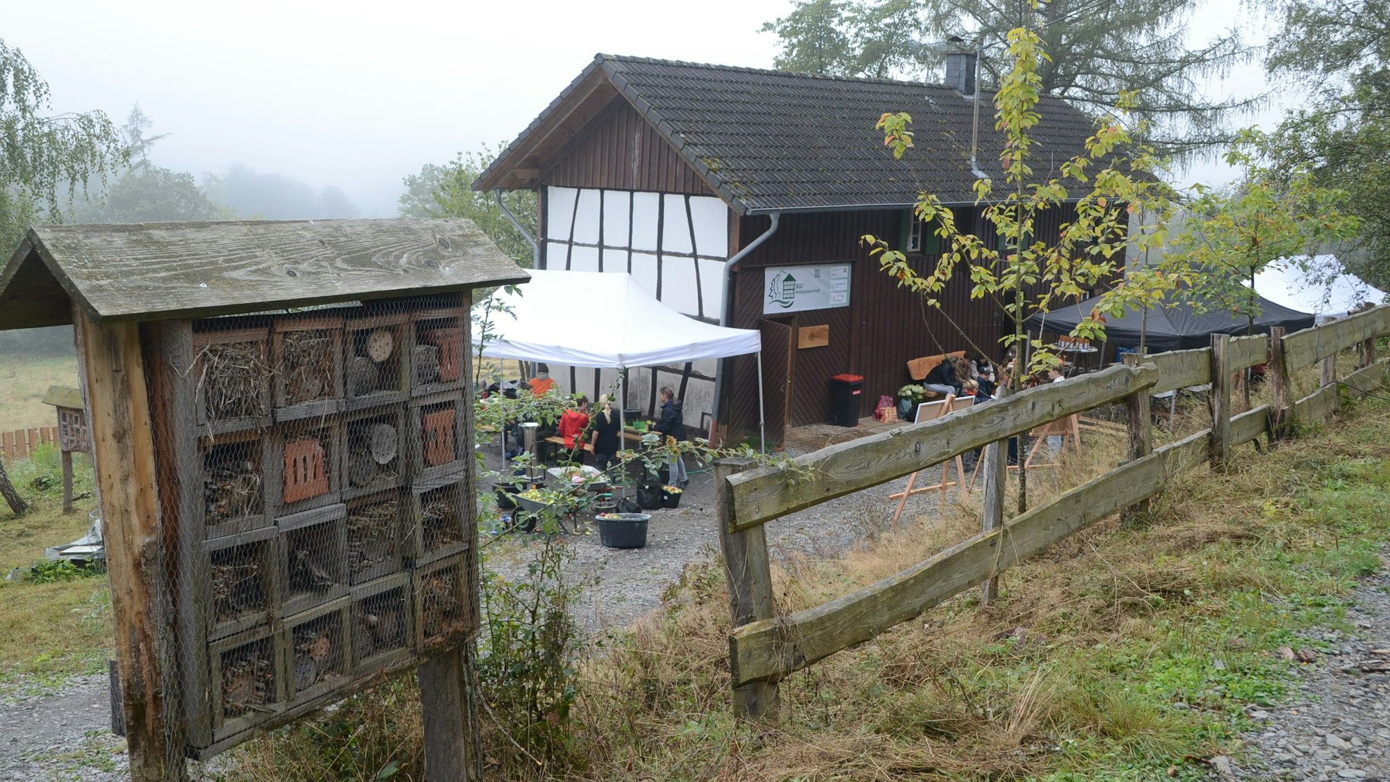 Ein Holzhaus mit Fachwerk an der Giebelwand. Im Vordergrund steht ein großes „Insektenhotel“.