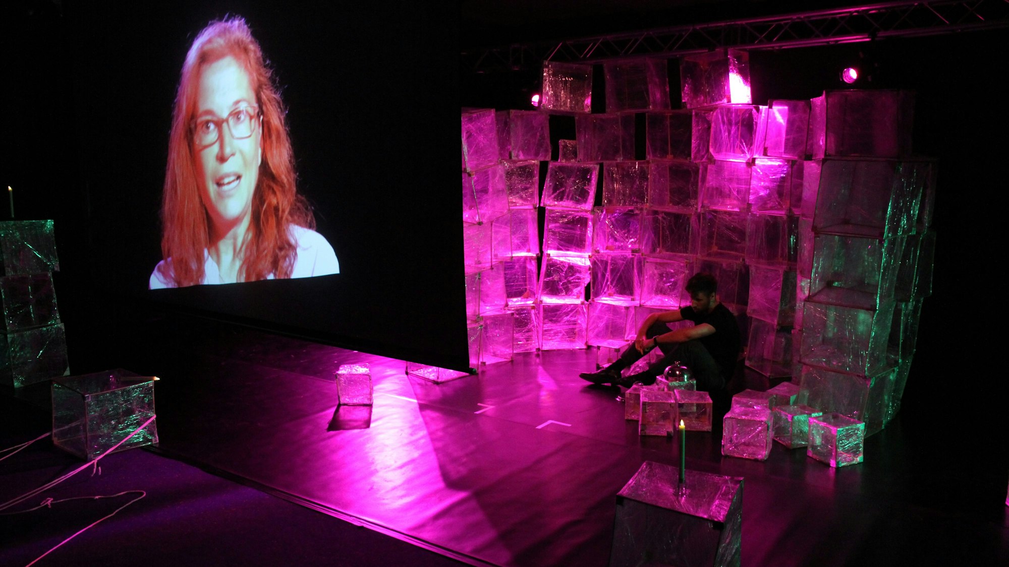 Ein Mann kauert vor Zellophanwürfeln auf einer Theaterbühne, links eine Frau auf einer Videoleinwand.