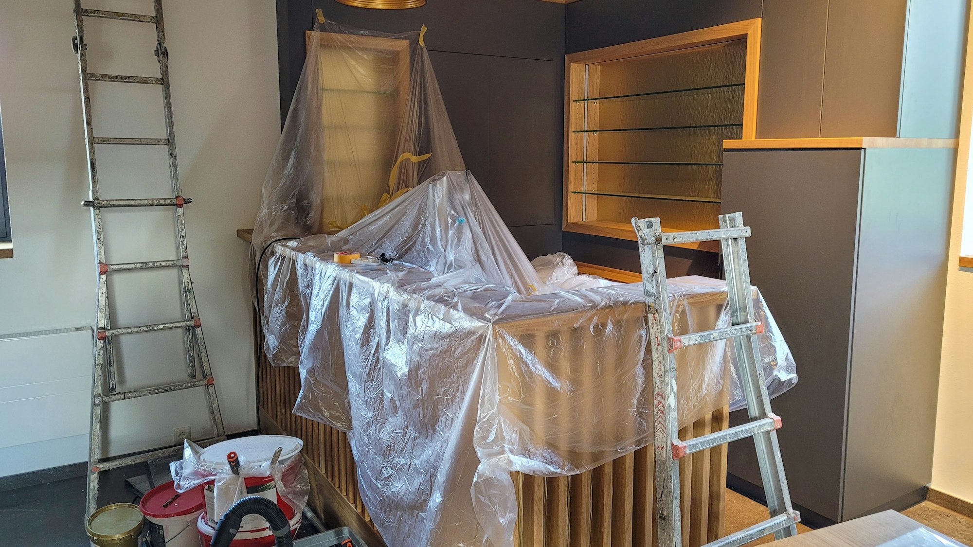 Unter durchsichtiger Malerfolie ist eine Holztheke zu erkennen, davor stehen Farbeimer und Pinsel, an der Wand lehnt eine Leiter.