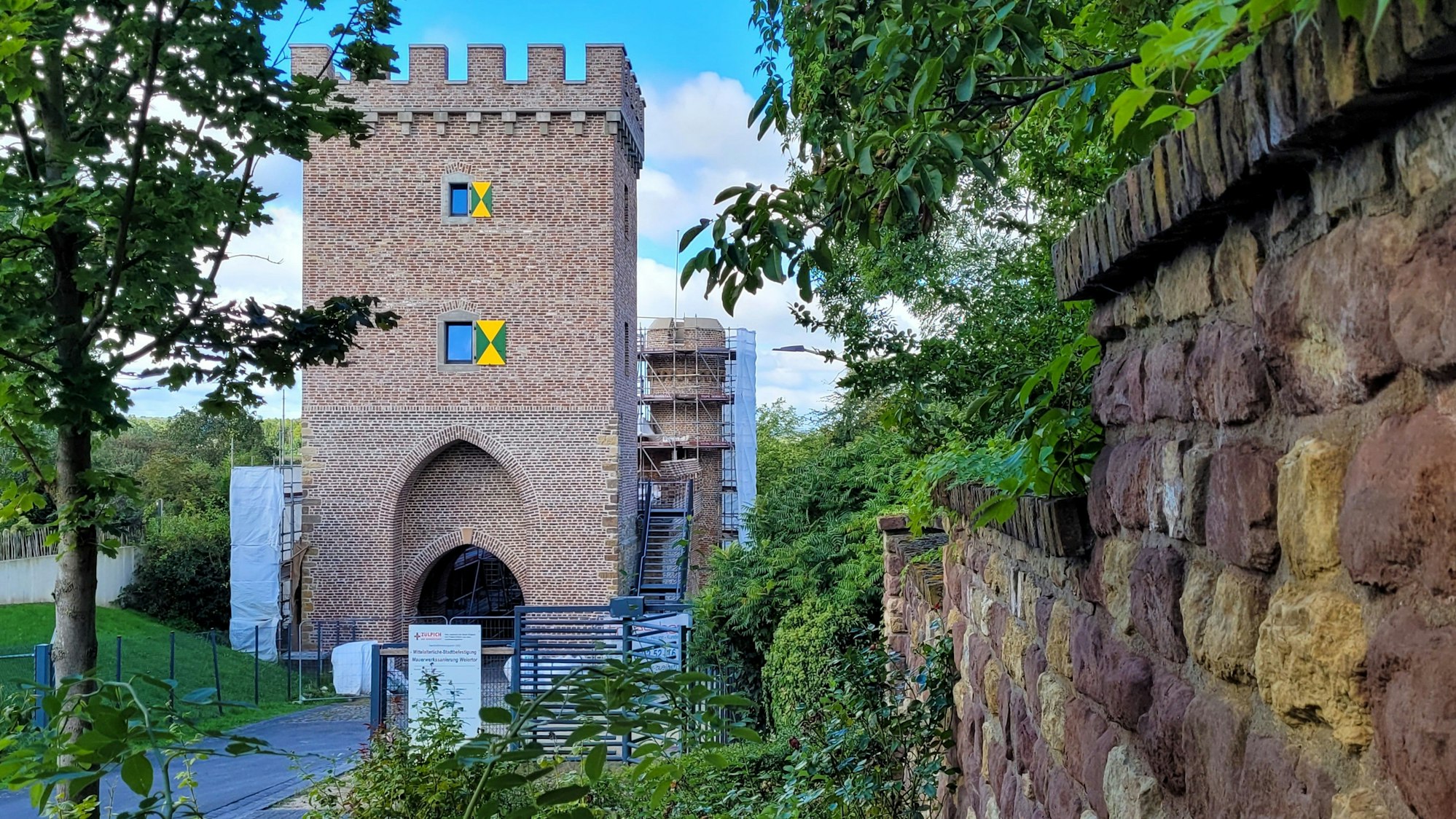 Hinter Büschen und Bäumen sieht man das neue Weiertor mit zwei Fenstern und grün-gelben Fensterläden.