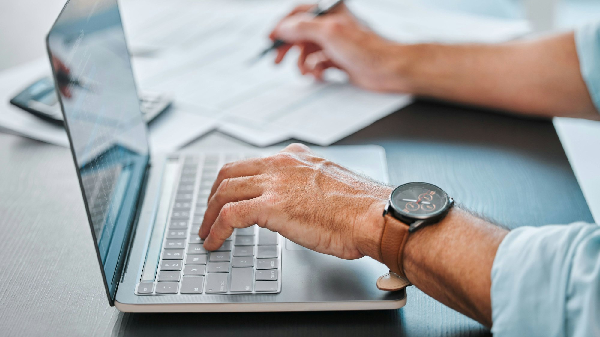 Zwei Hände auf einem Schreibtisch, eine ist auf der Tastatur eines Laptops, die andere schreibt etwas mit einem Stift auf Papier