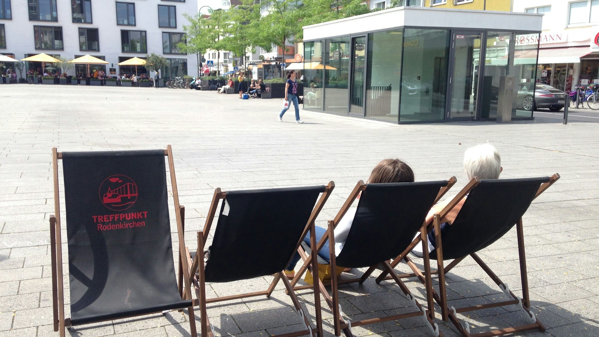 Zu sehen sind vier Liegestühle auf dem Maternusplatz in Rodenkirchen. In zwei von ihnen sitzen Personen.