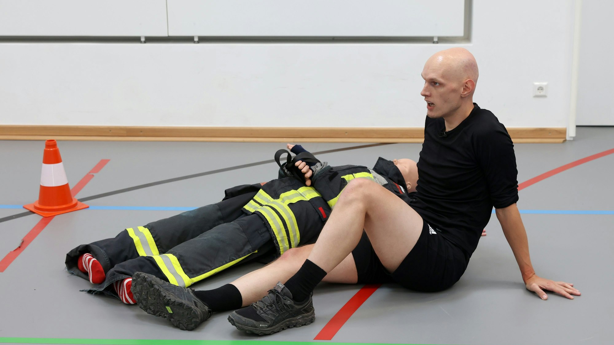 Florian Holler sitzt neben einem Dummy auf dem Boden einer Turnhalle.