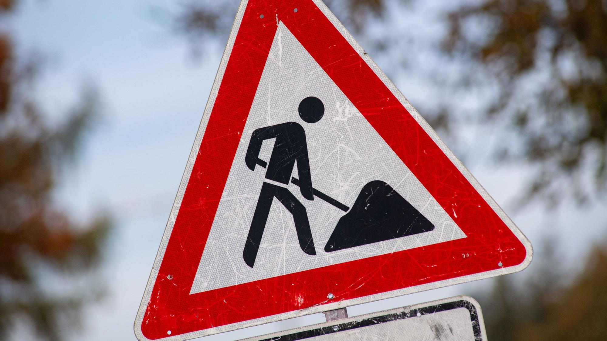 Auf dem Bild ist Ein zerkratztes Schild, Gefahrzeichen Arbeitsstelle (Zeichen 123, mit dem Piktogramm eines Bauarbeiters zur Beschilderung einer Baustelle zu sehen.