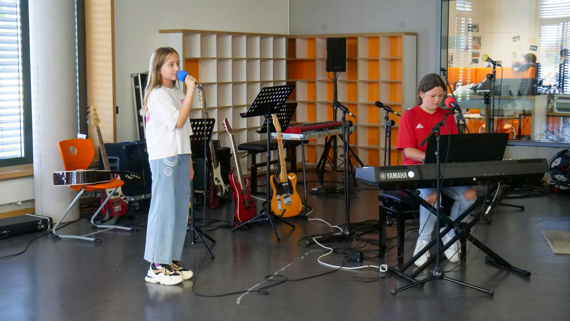 Ein Mädchen singt, ein anderes spielt Keyboard, im Hintergrund stehen Gitarren.
