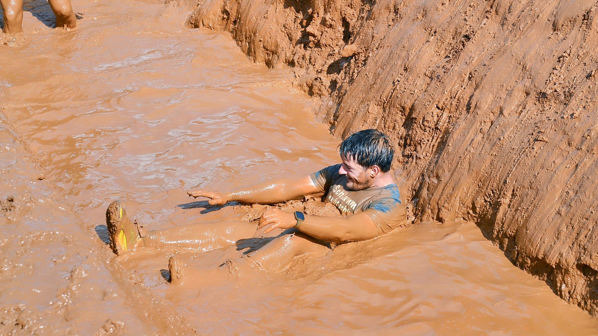 Ein Mann sitzt in verschlammtem Wasser, nachdem er ein Hindernis überquert hat. Er lacht.