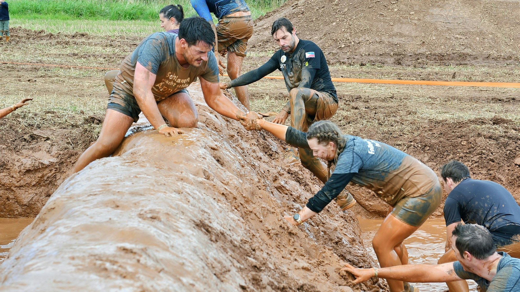 Mehrere Teilnehmerinnen und Teilnehmer überqueren einen verschlammten Damm. Ein Mann reicht einer Frau die Hand, um ihr zu helfen.
