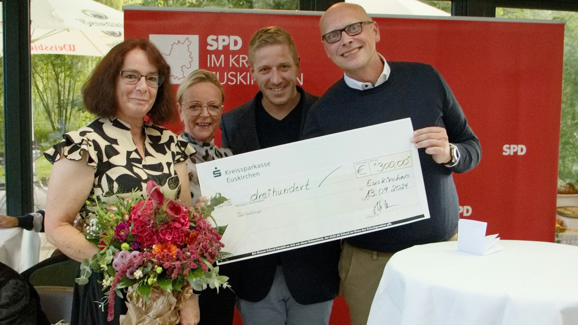 Eine Frau hält einen Blumenstrauß in der Hand, eine weitere Frau und zwei Männer neben ihr zeigen einen symbolischen Scheck.