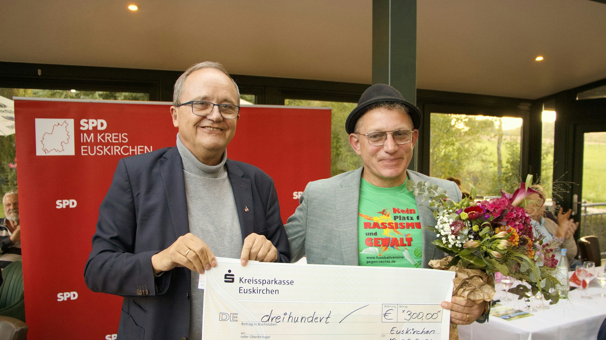 Die beiden Männer lächeln in die Kamera, sie halten den symbolischen Scheck fest.