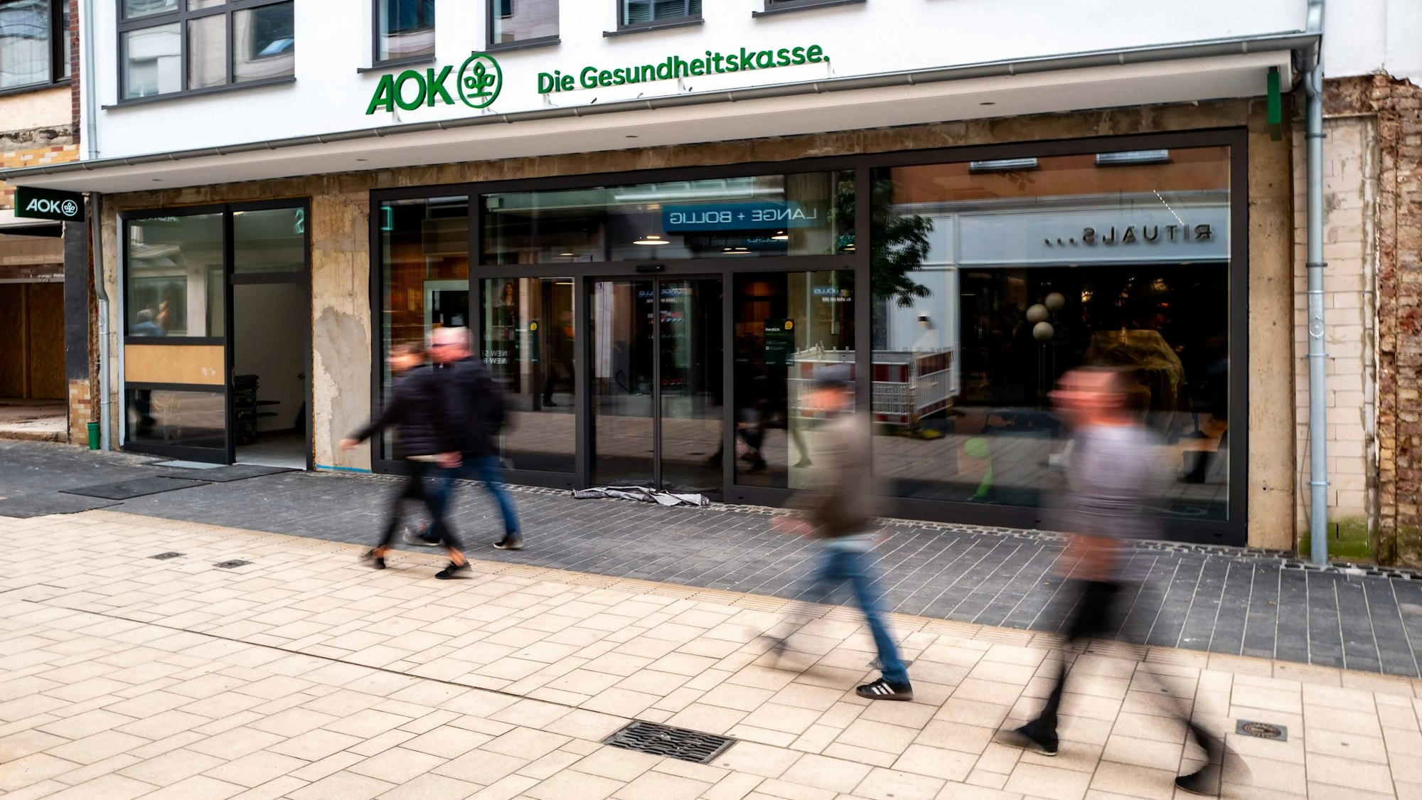 Über einem Eingang in der Euskirchener Innenstadt prangt der Schriftzug der AOK. Unscharf sind davor vier Menschen zu erkennen, die an der neuen Geschäftsstelle der Krankenkasse vorbeigehen.