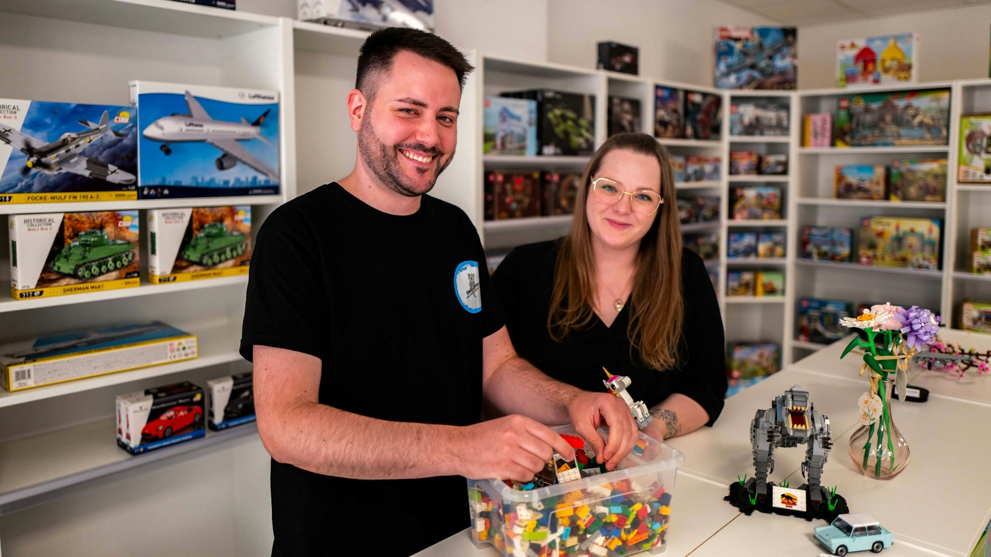 Kai Spessart und Lisa Wenkemann stehen in ihrem Geschäft für Klemmbausteine in Euskirchen. Vor ihnen auf dem Tresen steht eine Kiste mit bunten Bausteinen.