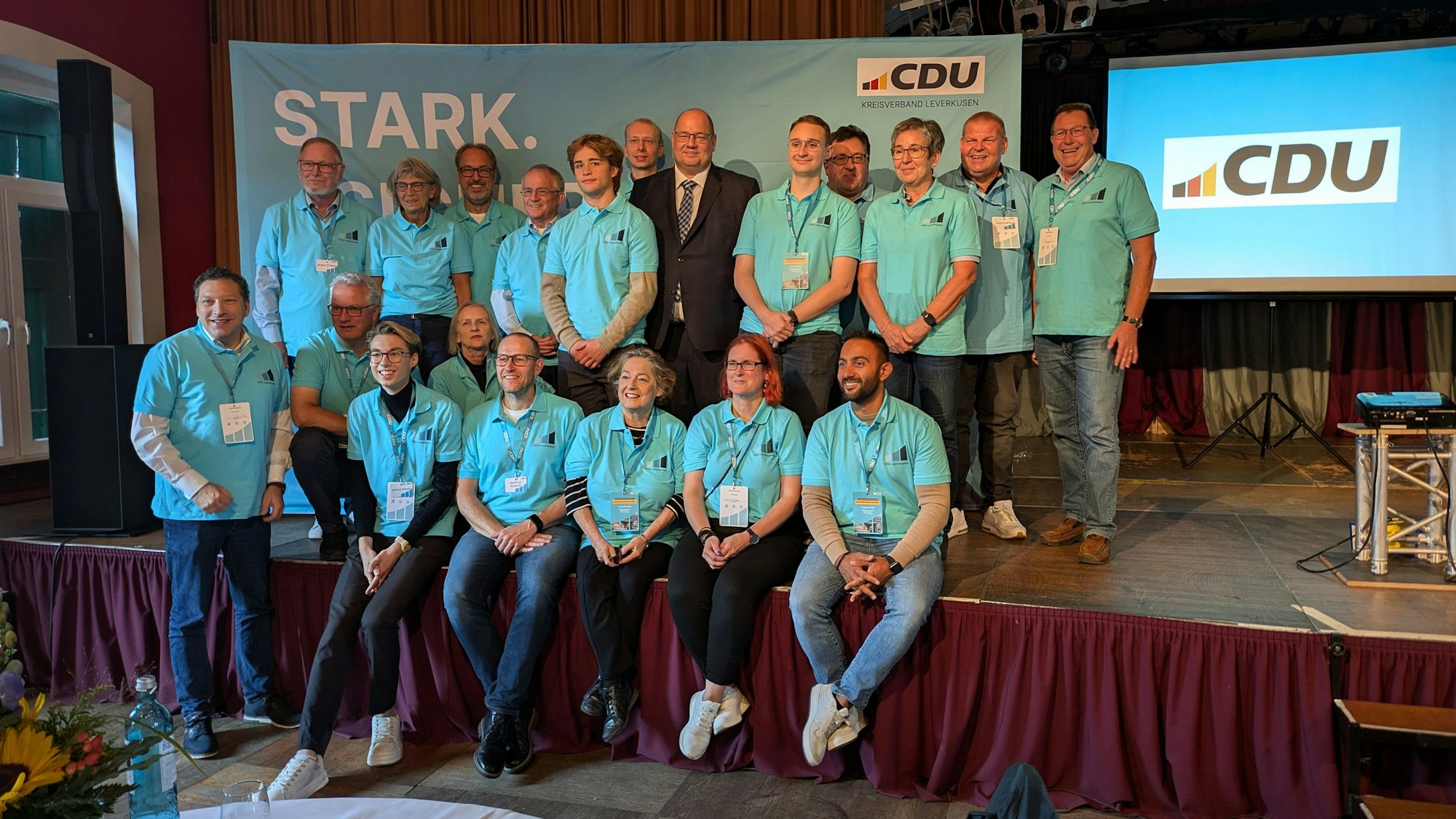 Eine Gruppe von Männern und Frauen in blauen T-Shirts umringt einen Mann im Anzug auf einer Bühne.