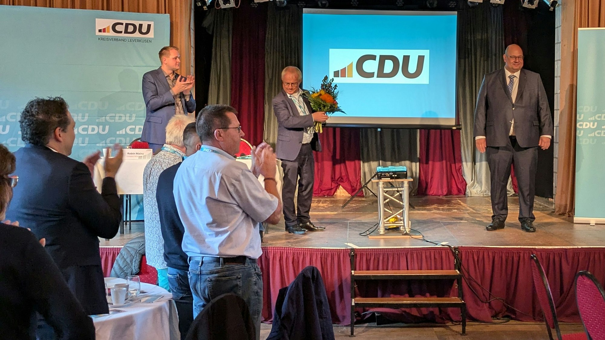 Männer applaudieren einem anderen Mann auf einer Bühne mit einem CDU-Plakat.