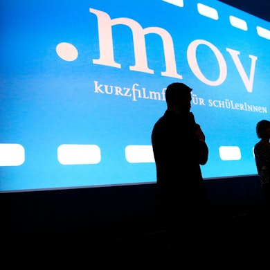 Zwei Moderatoren stehen vor einer Kinoleinwand.