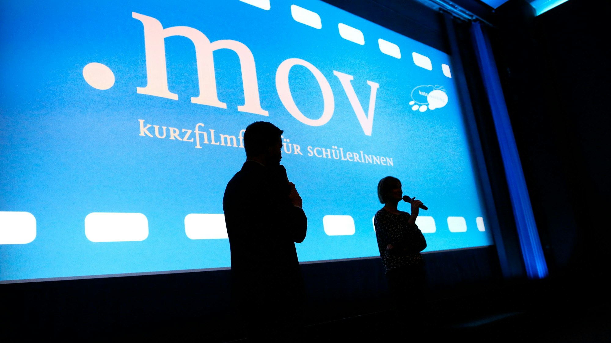 Zwei Moderatoren stehen vor einer Kinoleinwand.