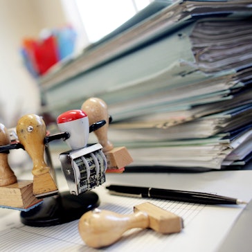 Ein Stempelkarussell steht neben einem Stapel Akten auf einem Schreibtisch in einer Behörde.