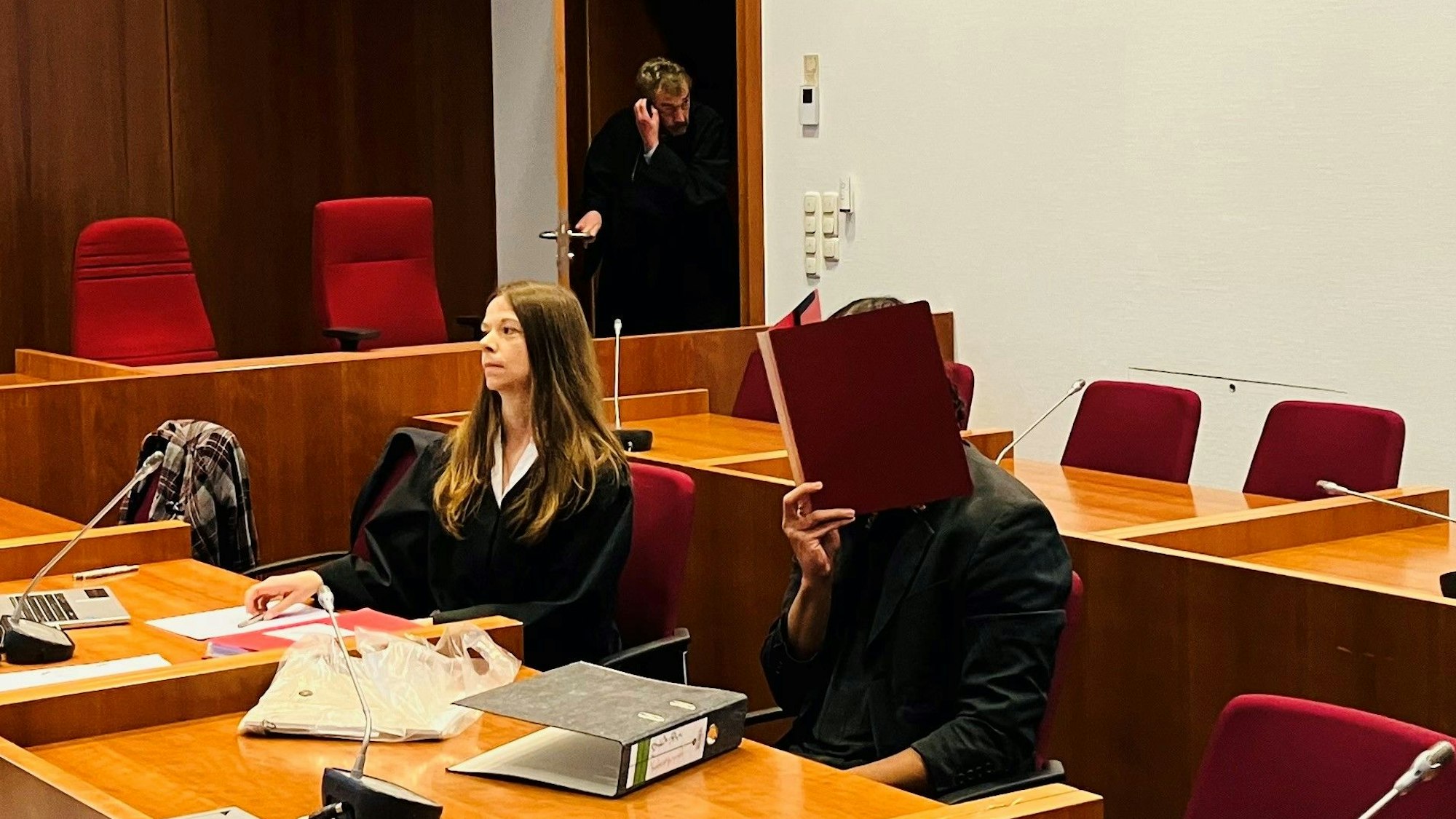Ein Mann in einem Gerichtssaal hält sich einen Aktenordner vor das Gesicht