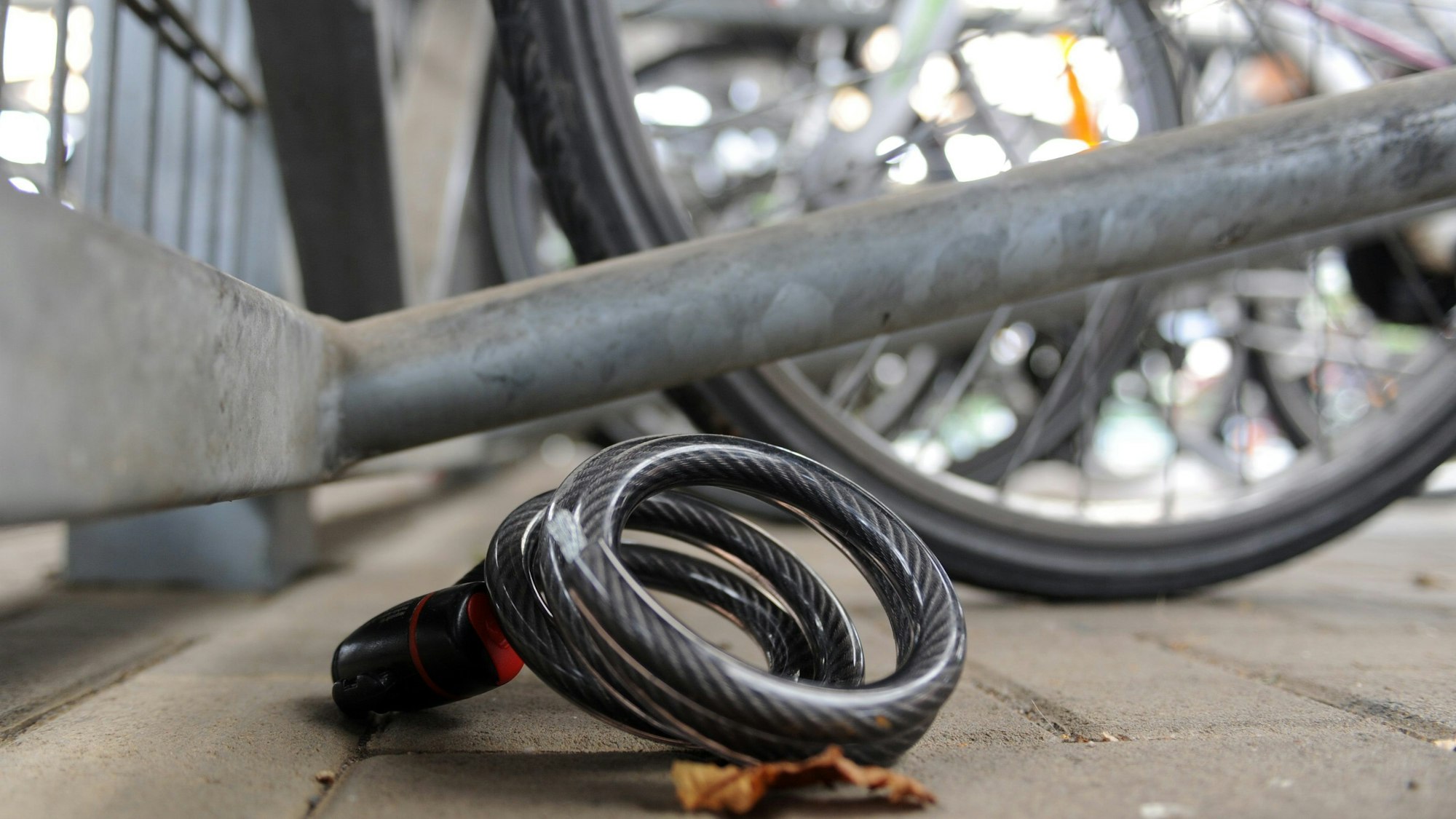 Auf dem Foto ist ein Fahrradschloss zu sehen, das auf dem Boden liegt, dahinter sind Fahrräder zu sehen.