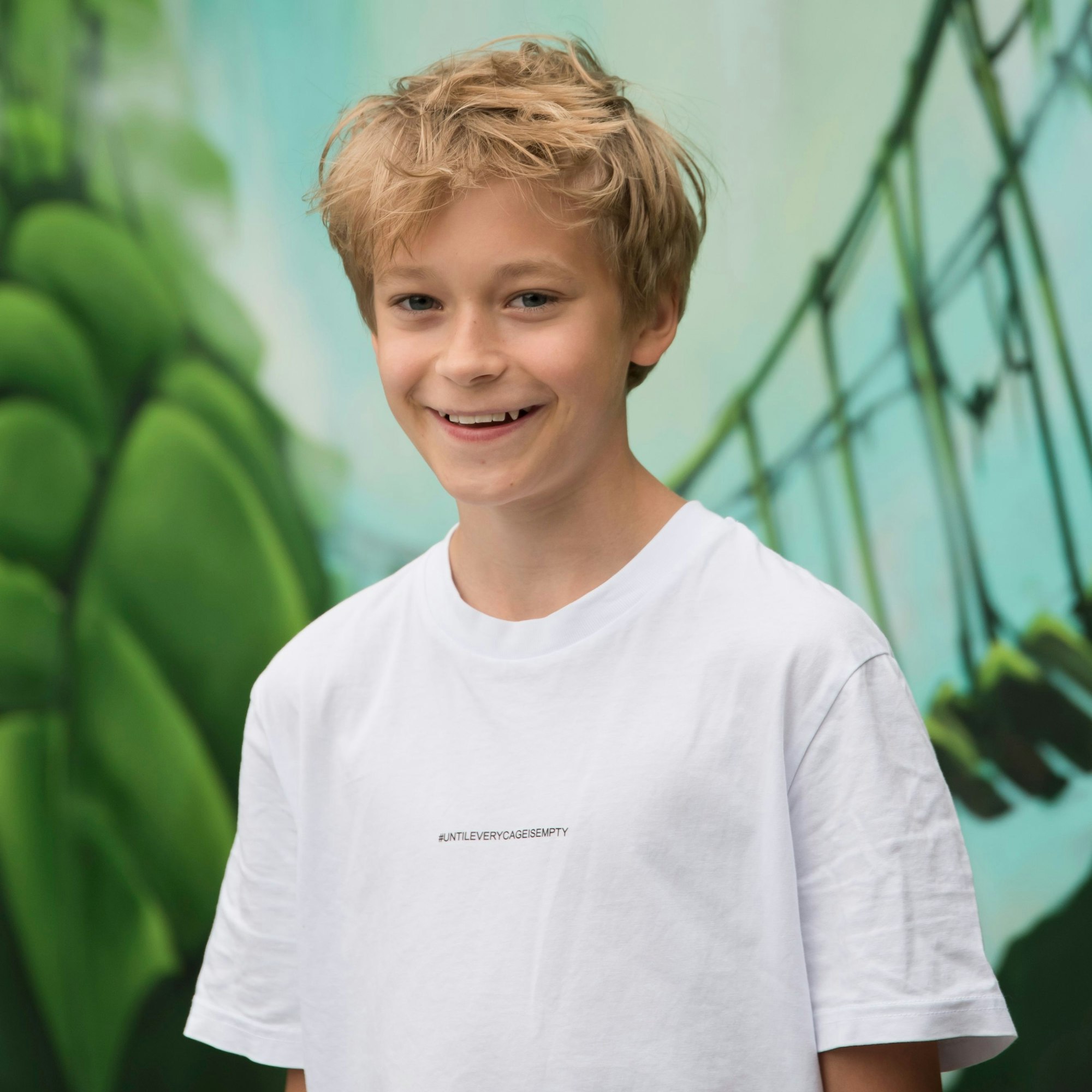 Henri steht vor einer Dschungel-Tapete ganz in grünen Tönen und lacht in die Kamera. Er trägt ein weißes T-Shirt und hat blondes kurzes Haar.
