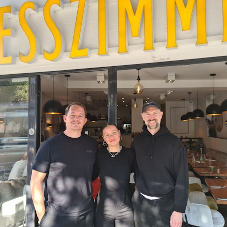 Zwei Männer umrahmen eine Frau, alle tragen schwarze Oberteile, sie stehen vor der geöffneten großen Glastür zu einem freundlich eingerichteten Gastraum.