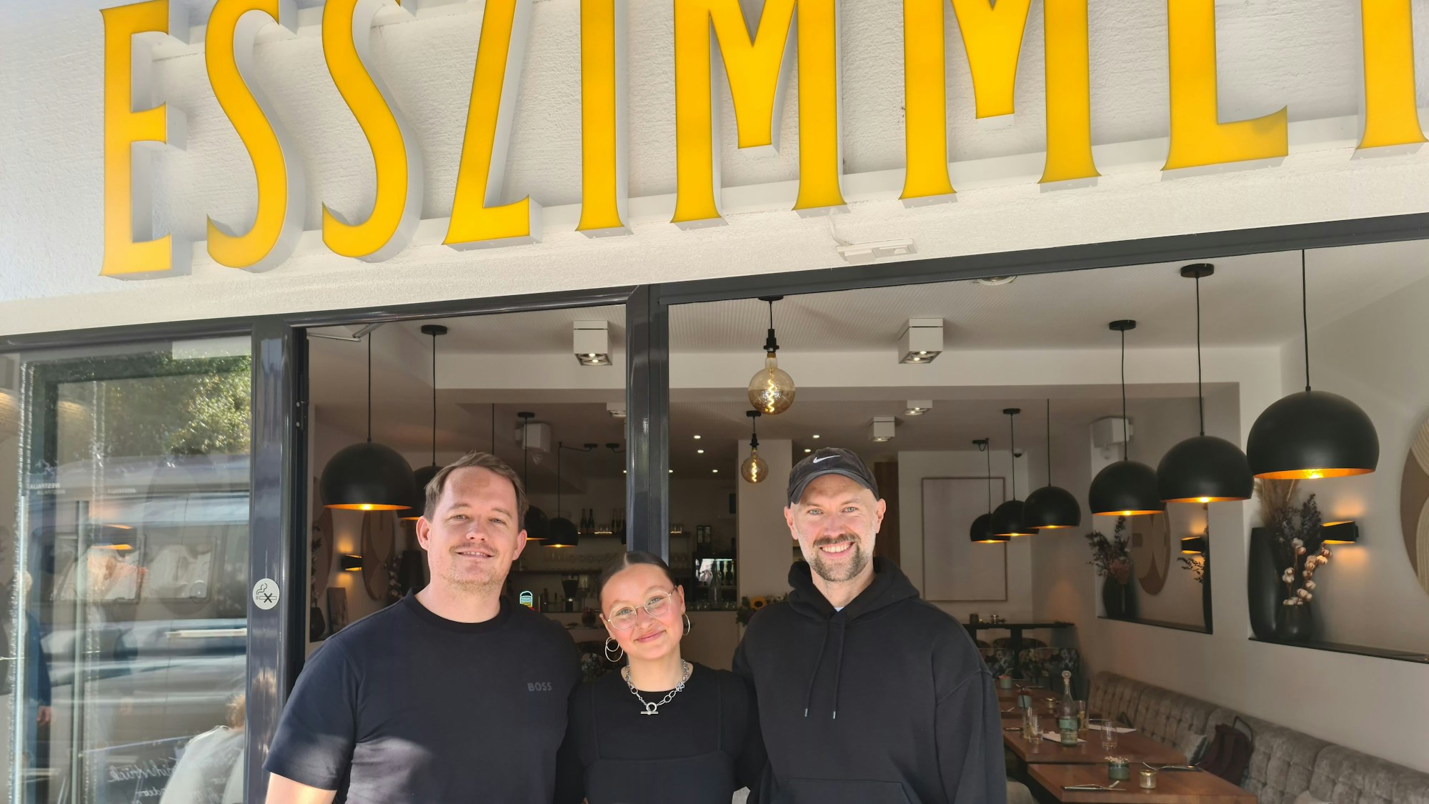 Zwei Männer umrahmen eine Frau, alle tragen schwarze Oberteile, sie stehen vor der geöffneten großen Glastür zu einem freundlich eingerichteten Gastraum.