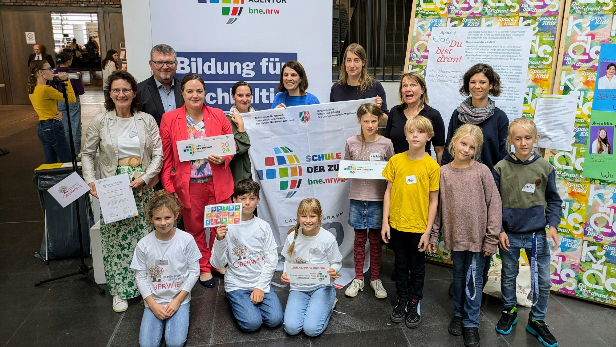 Grundschüler und Erwachsene stehen in einer Gruppe zusammen, sie zeigen gemeinsam ein Banner für das Programm „Schule der Zukunft“.