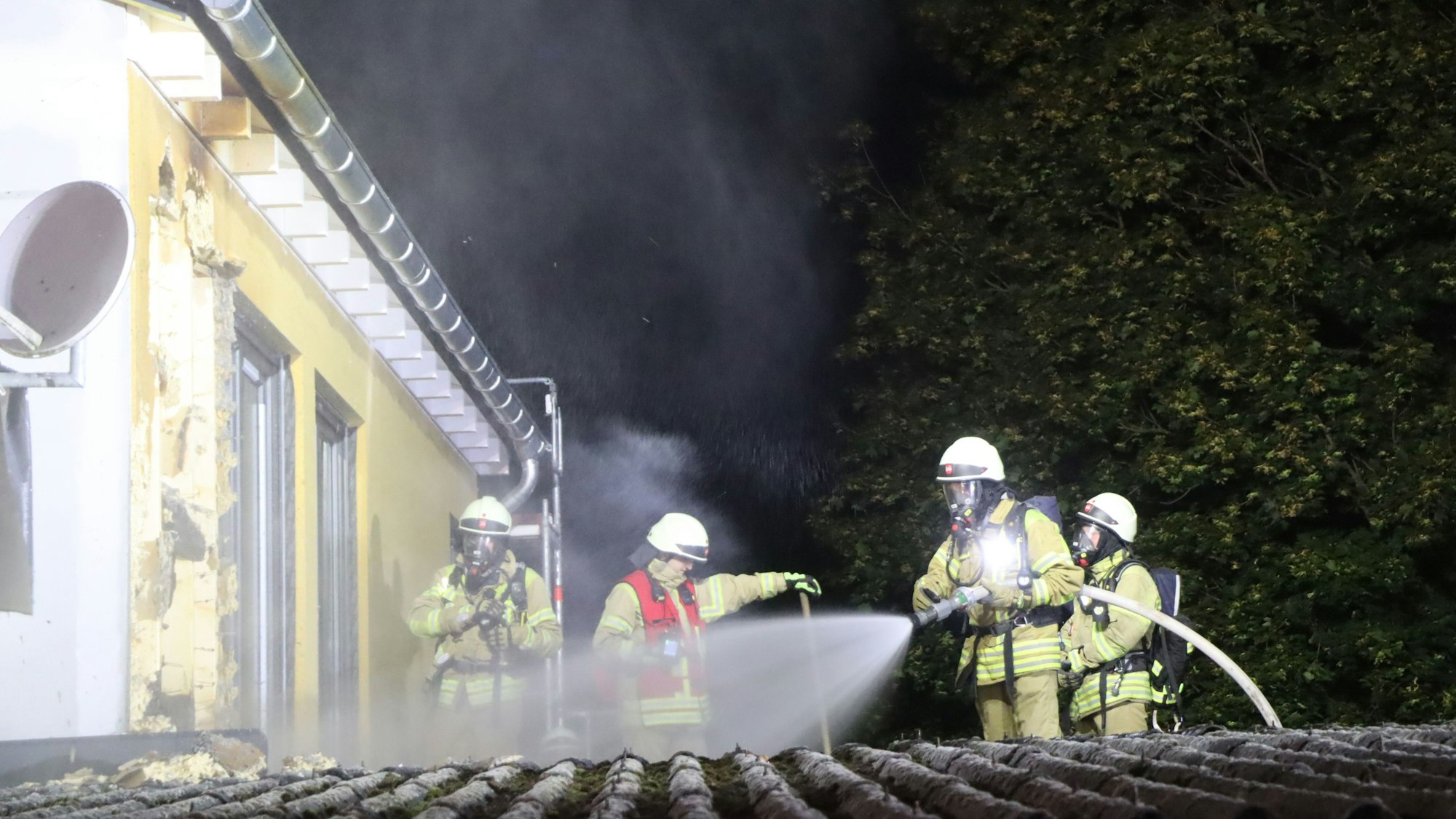 Feuerwehrleute stehen auf einem Dach und sprühen mit einem Wasserschlauch auf eine gelbe Fassade.
