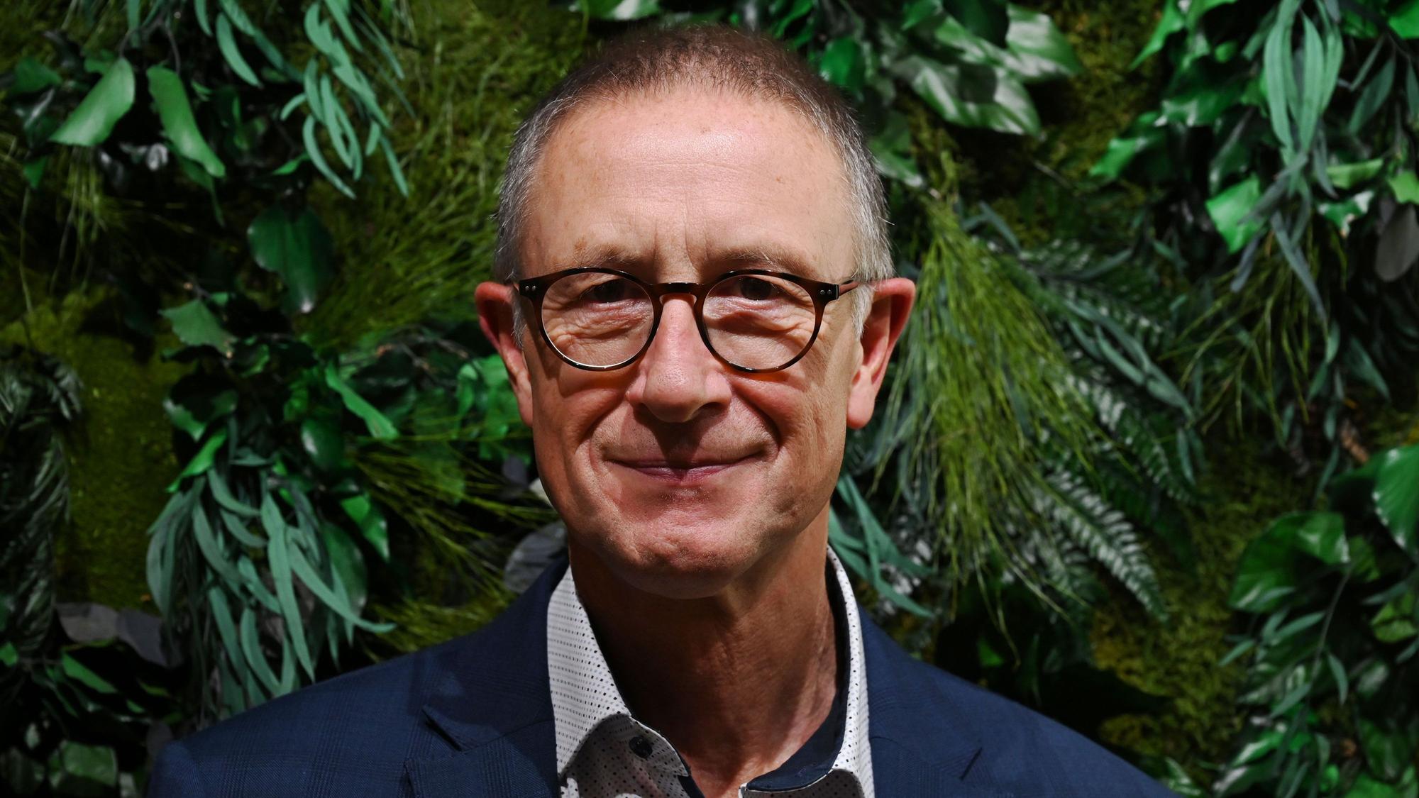 Helmut Schneider, Regionaldirektor der AOK Rheinland/Hamburg, steht vor einer Wand mit grünen Pflanzen.