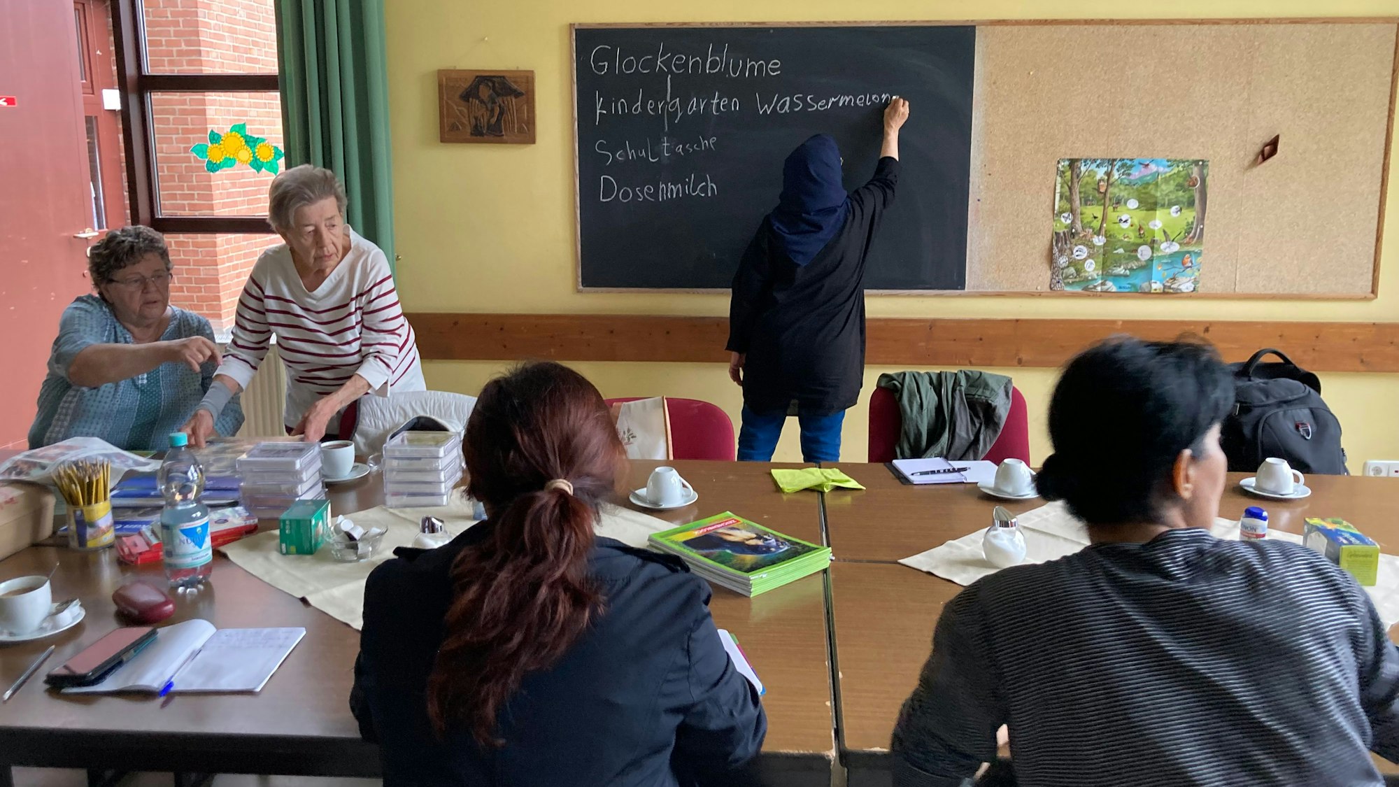 Eine ältere Frau zeigt einer anderen älteren Frau etwas, das sich außerhalb des Bildrandes befindet, während eine Frau im Hidschāb das Wort Wassermelone an eine Tafel schreibt.