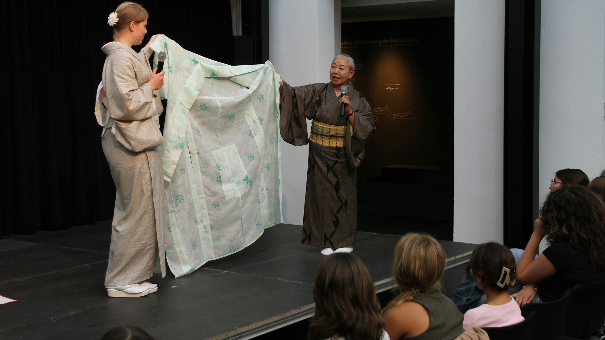 Zwei Frauen zeigen auf einer Bühne einen aufgefalteten Kimono