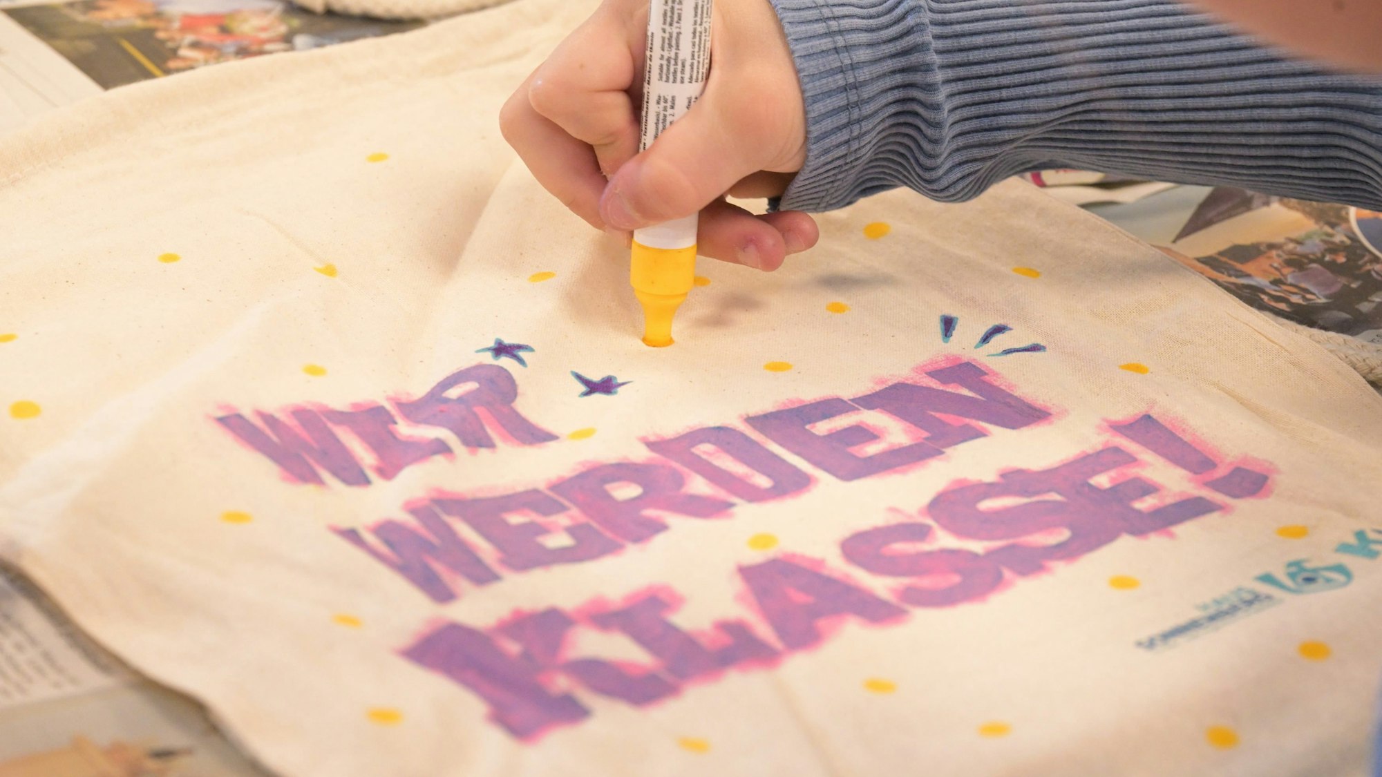 Ein Kind bemalt ein Stück Stoff mit dem Schriftzug: "Wir werden Klasse!"