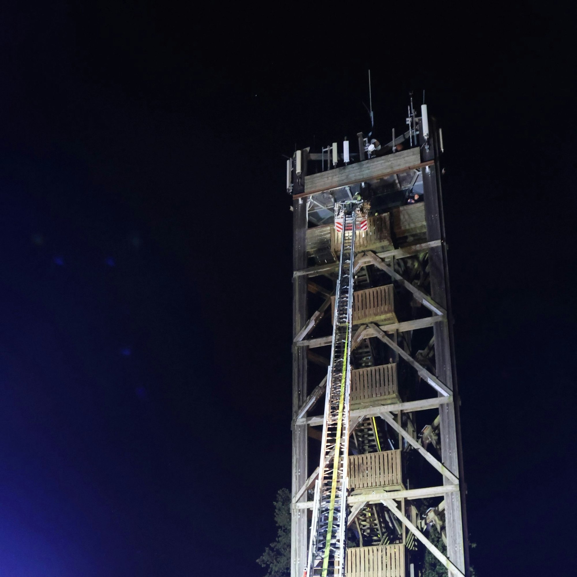 Vor dunklem Nachthimmel ist ein Teil des Aussichtsturms Nümbrecht zu erkennen, der Korb einer Feuerwehr-Drehleiter ist an dem Turm angelegt.