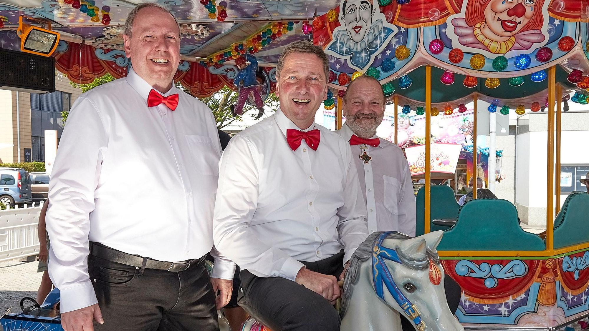 Joachim Vossel, Werner Echtner und Johann Manfred Klöcker haben sich für das Foto auf einem Kinderkarussell platziert. Sie tragen weiße Hemden und rote Fliegen.