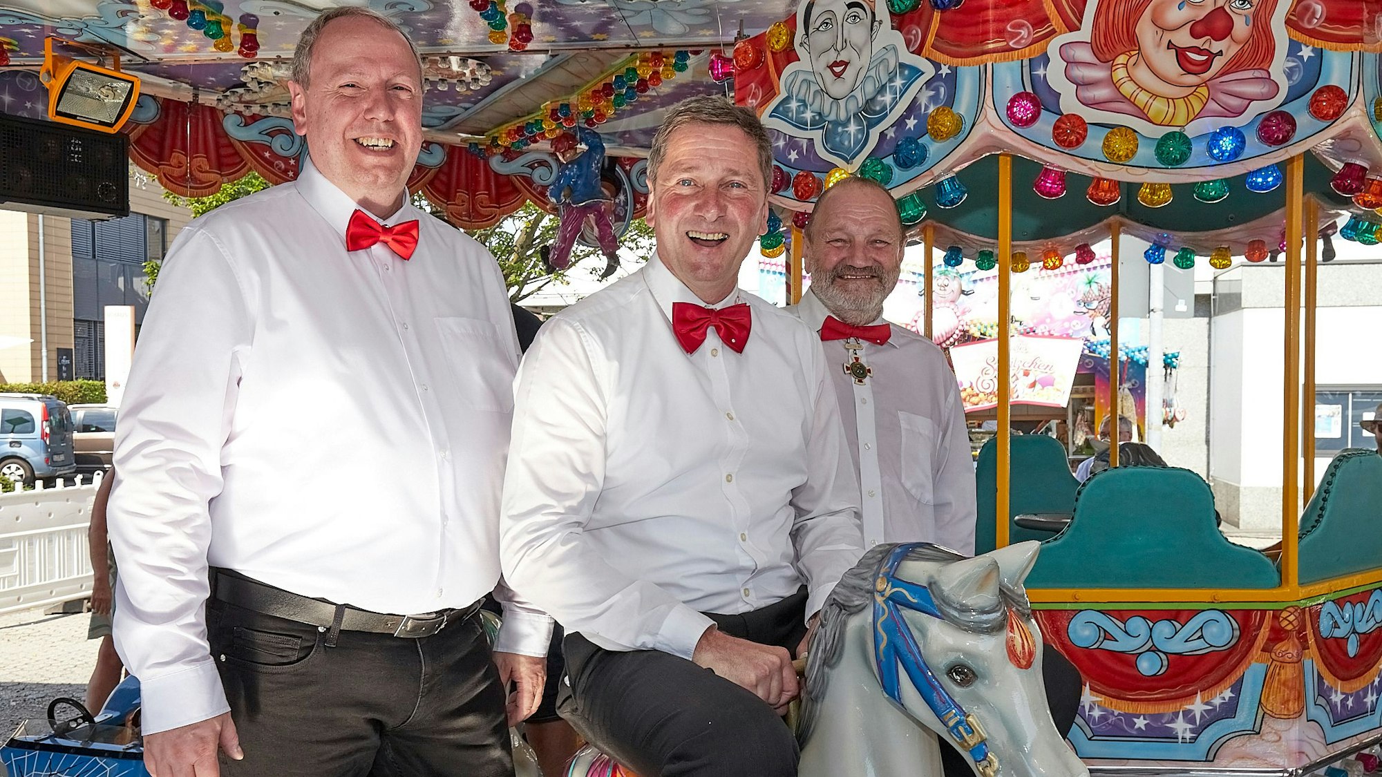 Joachim Vossel, Werner Echtner und Johann Manfred Klöcker haben sich für das Foto auf einem Kinderkarussell platziert. Sie tragen weiße Hemden und rote Fliegen.