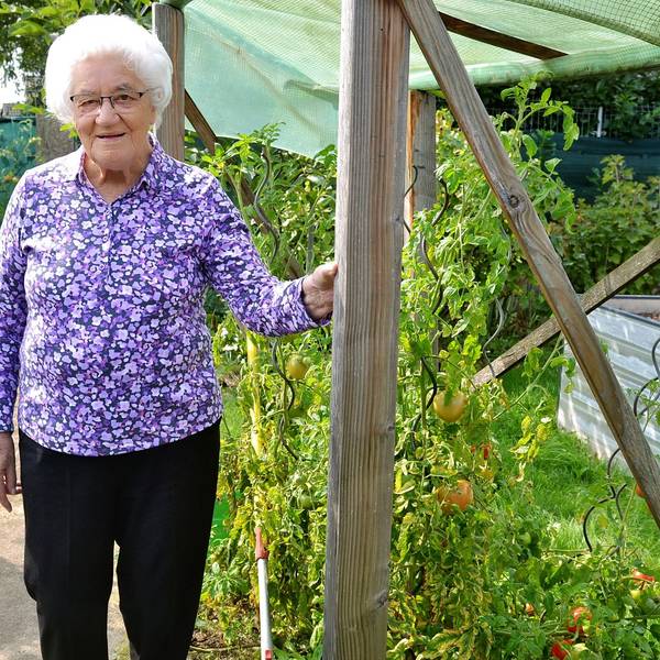 Eine ältere Frau in Bluse mit Blumenmuster steht in einem Garten und fasst mit ihrer linken Hand an einen Holzpfahl.