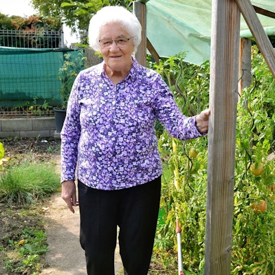 Eine ältere Frau in Bluse mit Blumenmuster steht in einem Garten und fasst mit ihrer linken Hand an einen Holzpfahl.