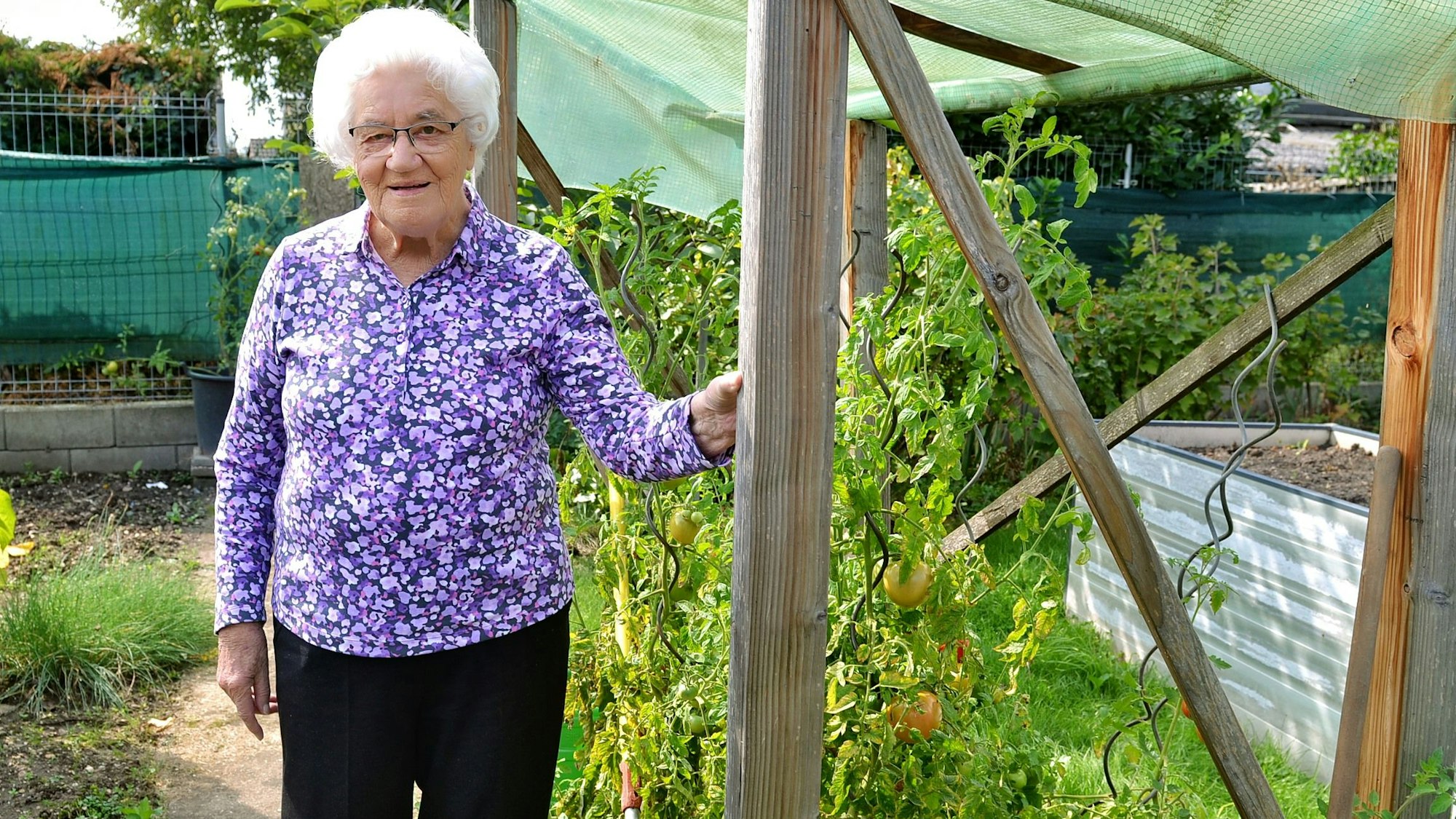 Eine ältere Frau in Bluse mit Blumenmuster steht in einem Garten und fasst mit ihrer linken Hand an einen Holzpfahl.
