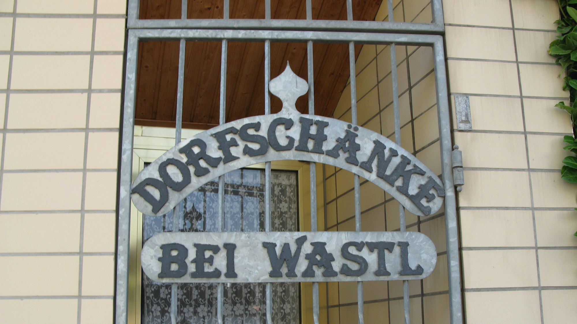 Ein Schild mit der Aufschrift „Dorfschänke Bei Wastl“ auf einem Metallgitter vor einer Tür.
