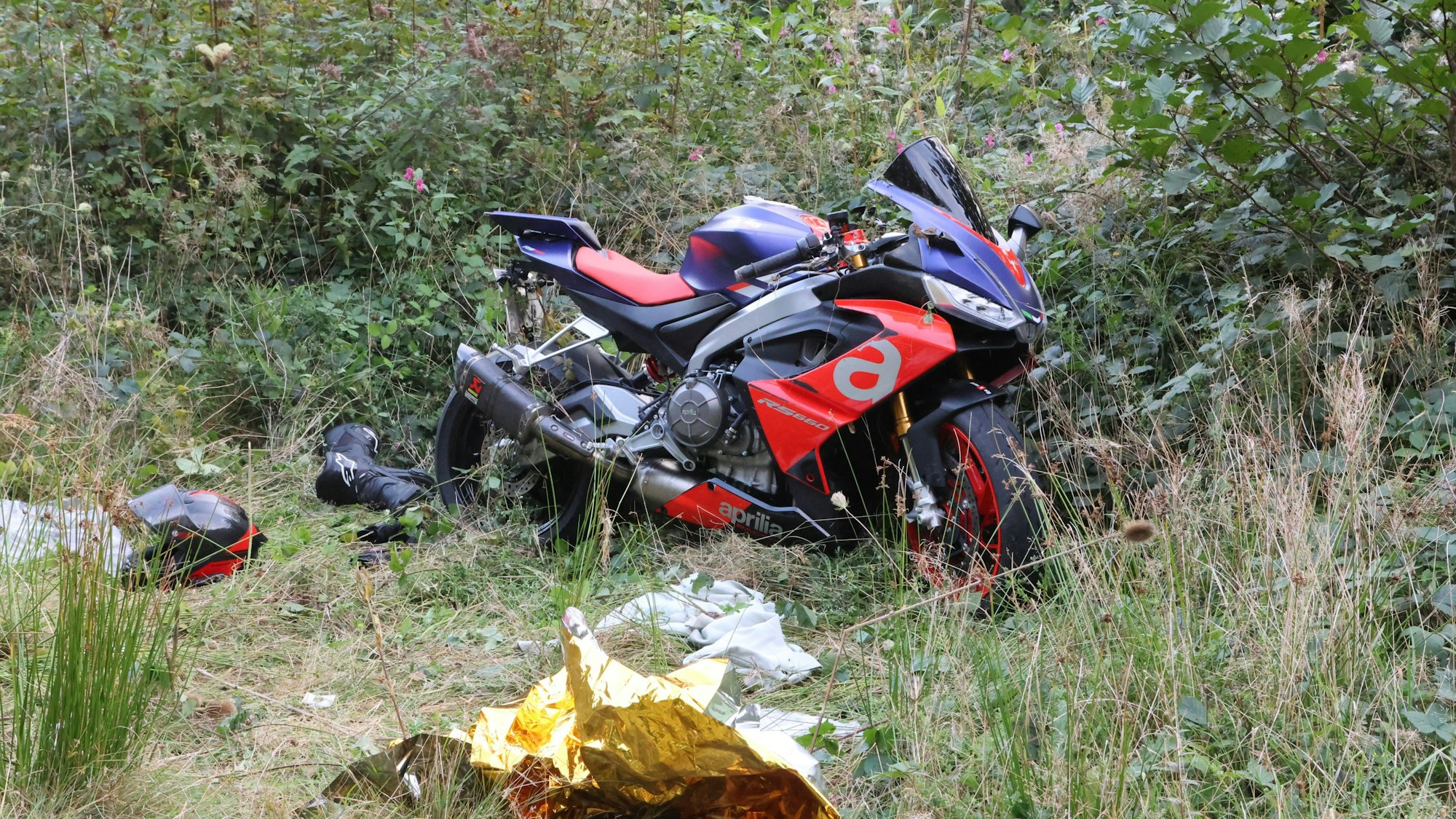 Ein Motorrad steht zwischen Büschen neben einer Straße, Rettungsdecken am Boden.