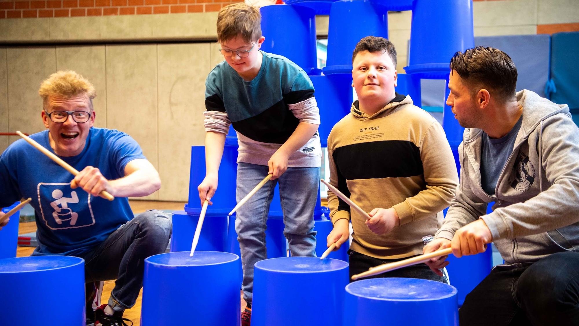 Das Bild zeigt zwei Schüler, die auf blaue Eimer eintrommeln. Zwei Lehrer unterstützen sie dabei.