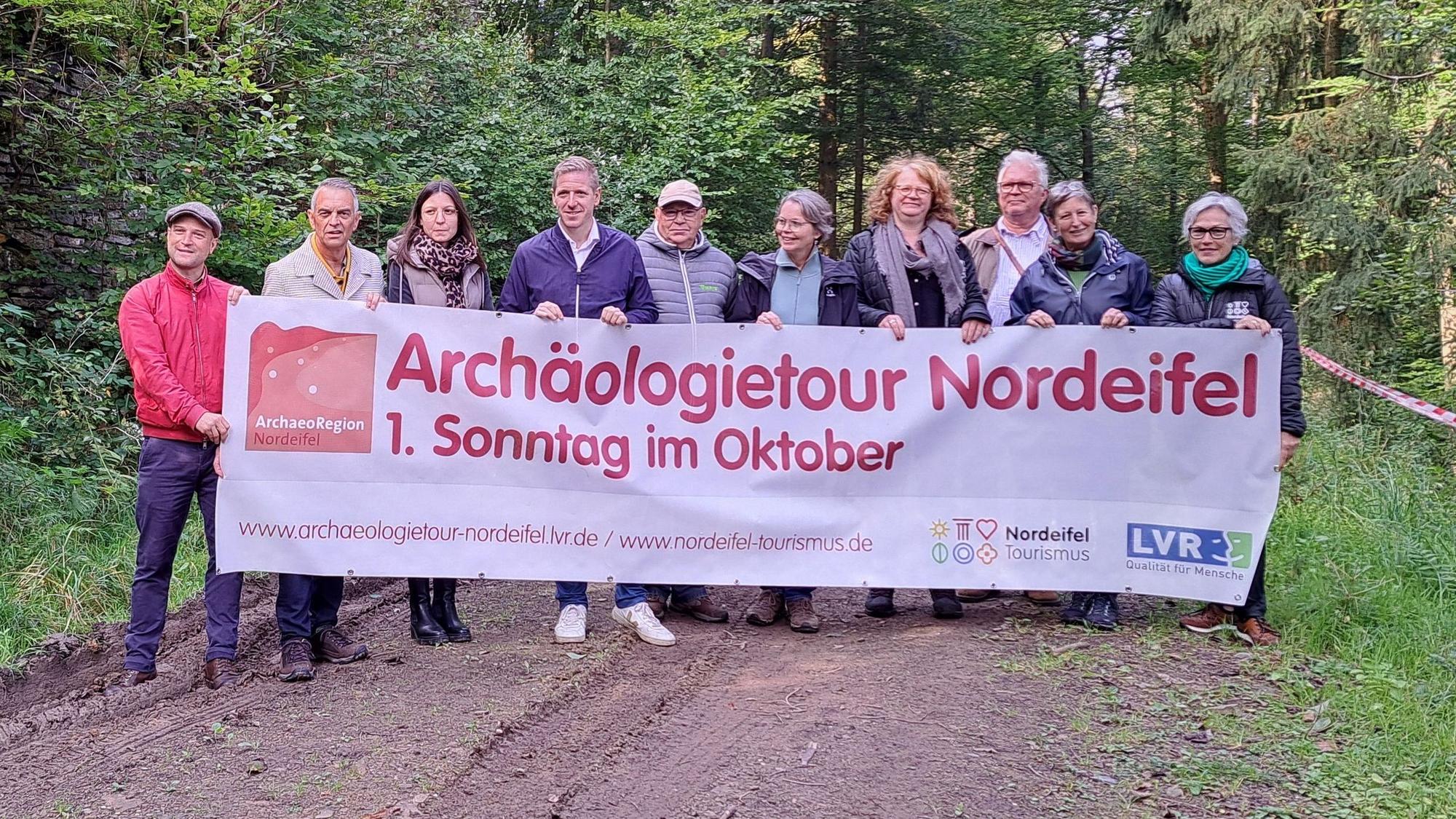 Eine Gruppe Menschen steht im Wald und hält ein Transparent, das für die Archäologietage wirbt.
